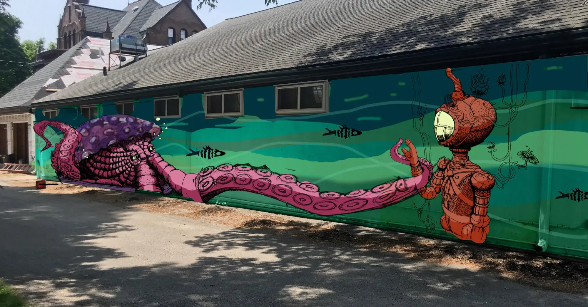 A long mural on a building wall: a huge pink octopus with a purple mantle reaches one tentacle across bands of green and teal painted water toward a clay-red ornamented diver, whose open hand meets it at fingertip range beneath a glowing oval faceplate.