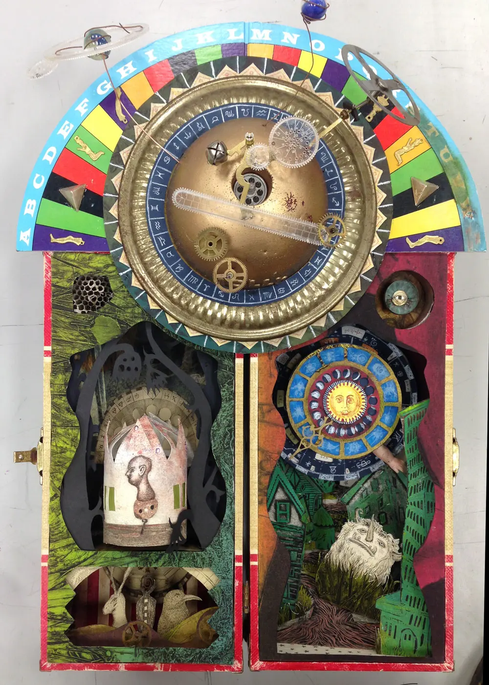 An assemblage almanach in a found doll case. A gilt dome with an orrery of clock parts and marbles crowns two chambers, one green and lit from within, one red with a sun face and a steep city street.