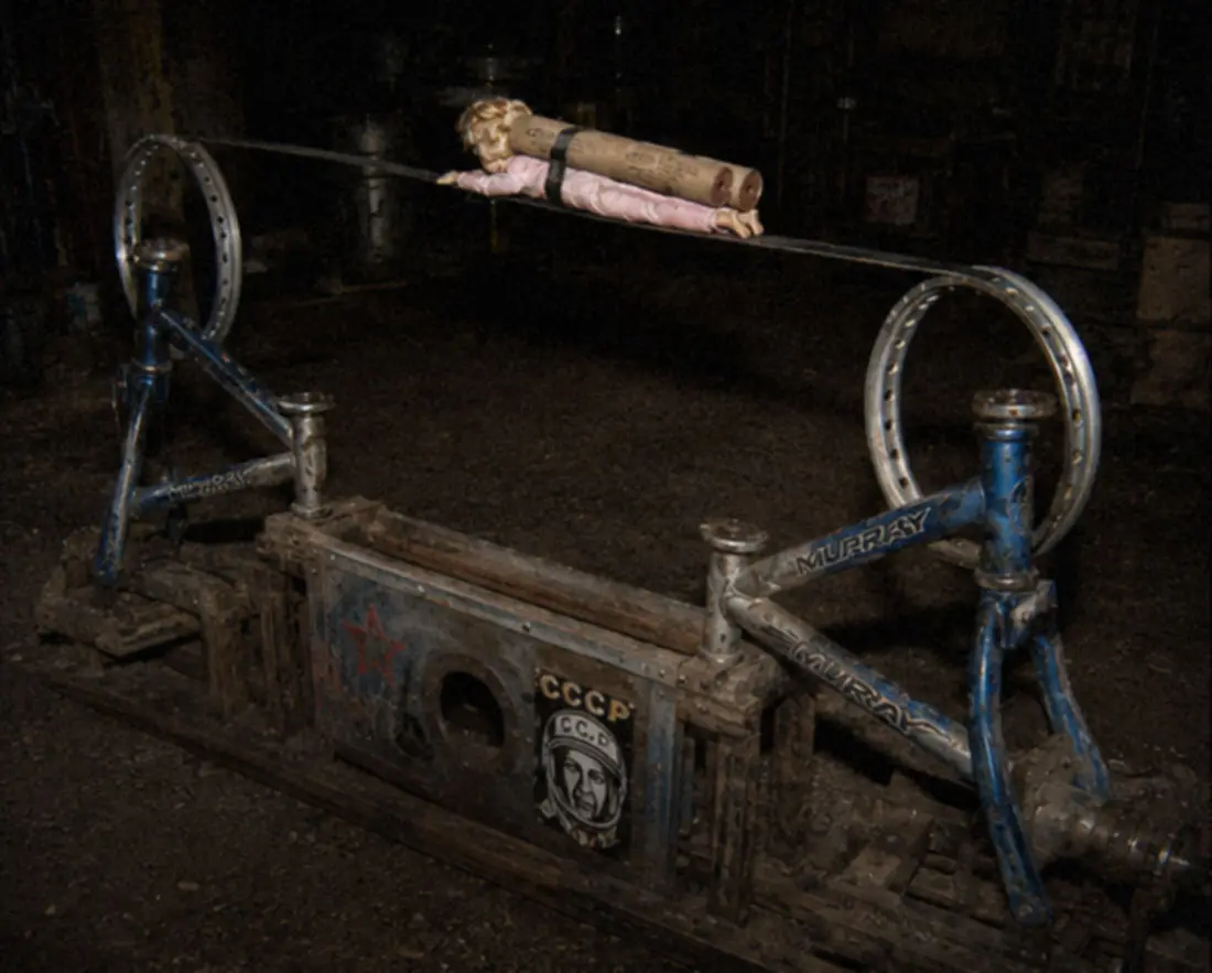 The launch rig in a dark shed: two blue Murray bicycle frames hold a pair of wheel-rim loops joined by a steel track, a doll in pink strapped face-down to cardboard-tube rockets on the rail, and a red star, CCCP lettering, and a stenciled cosmonaut portrait on the base.