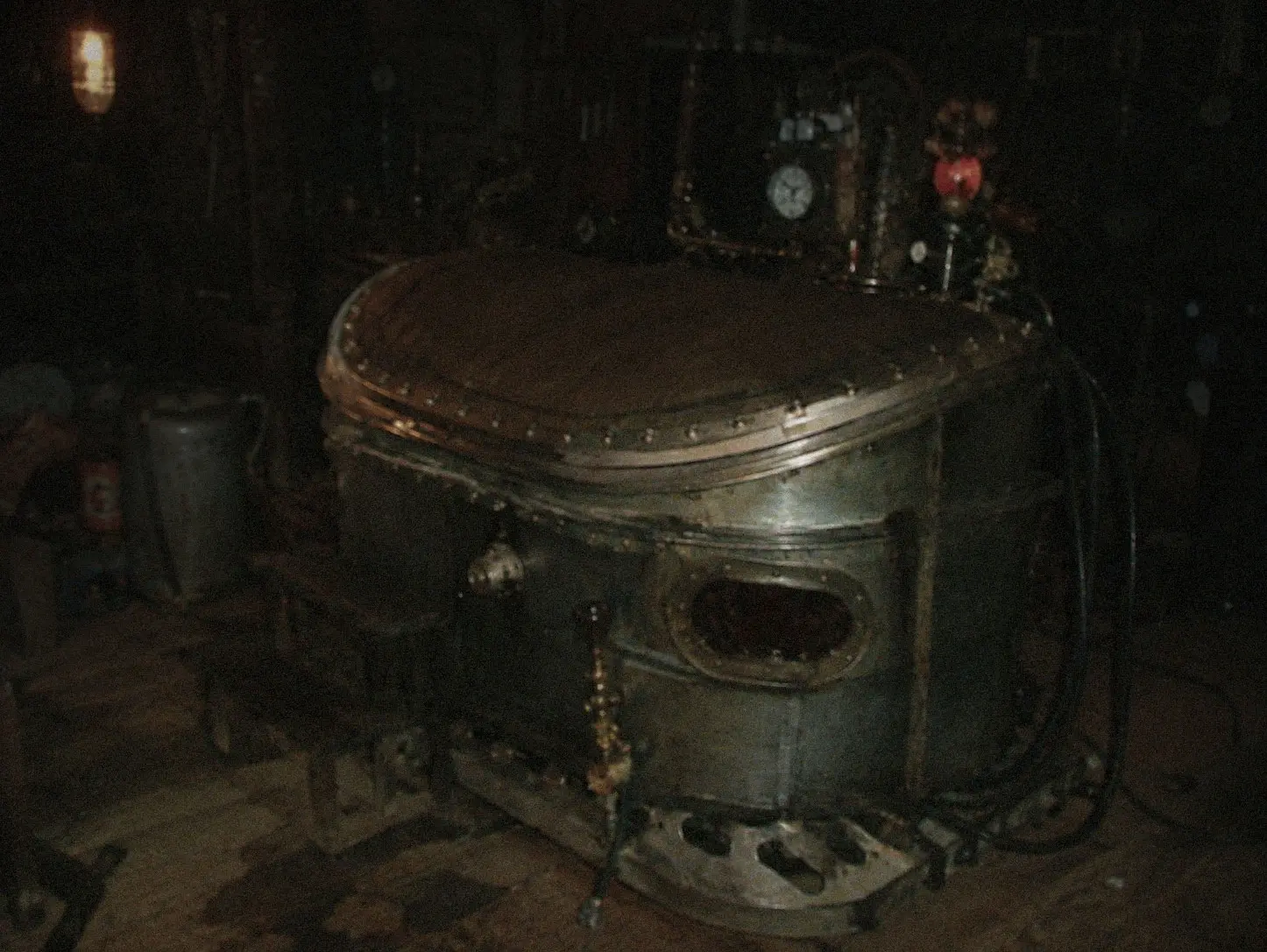 The chamber in a dark workshop: a galvanized oval tank on skids, a riveted wooden lid, a small porthole in the flank, and a panel above it crowded with a clock, gauges, and brass fittings.