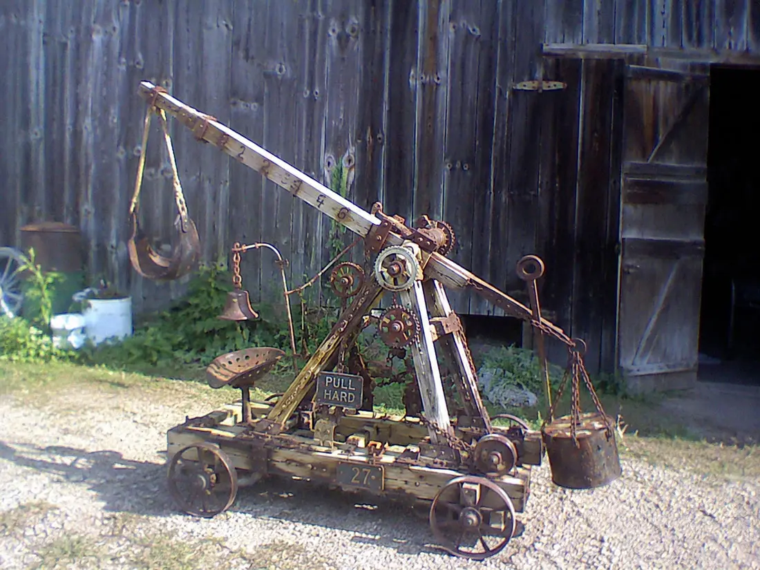 The trebuchet in gravel before a weathered gray barn: a numbered wooden throwing arm raised with a leather sling hung from its tip, a rusted counterweight bucket on chains, a tractor seat, a bell, gears and pulleys, a PULL HARD sign, and a 27 plate on the carriage.