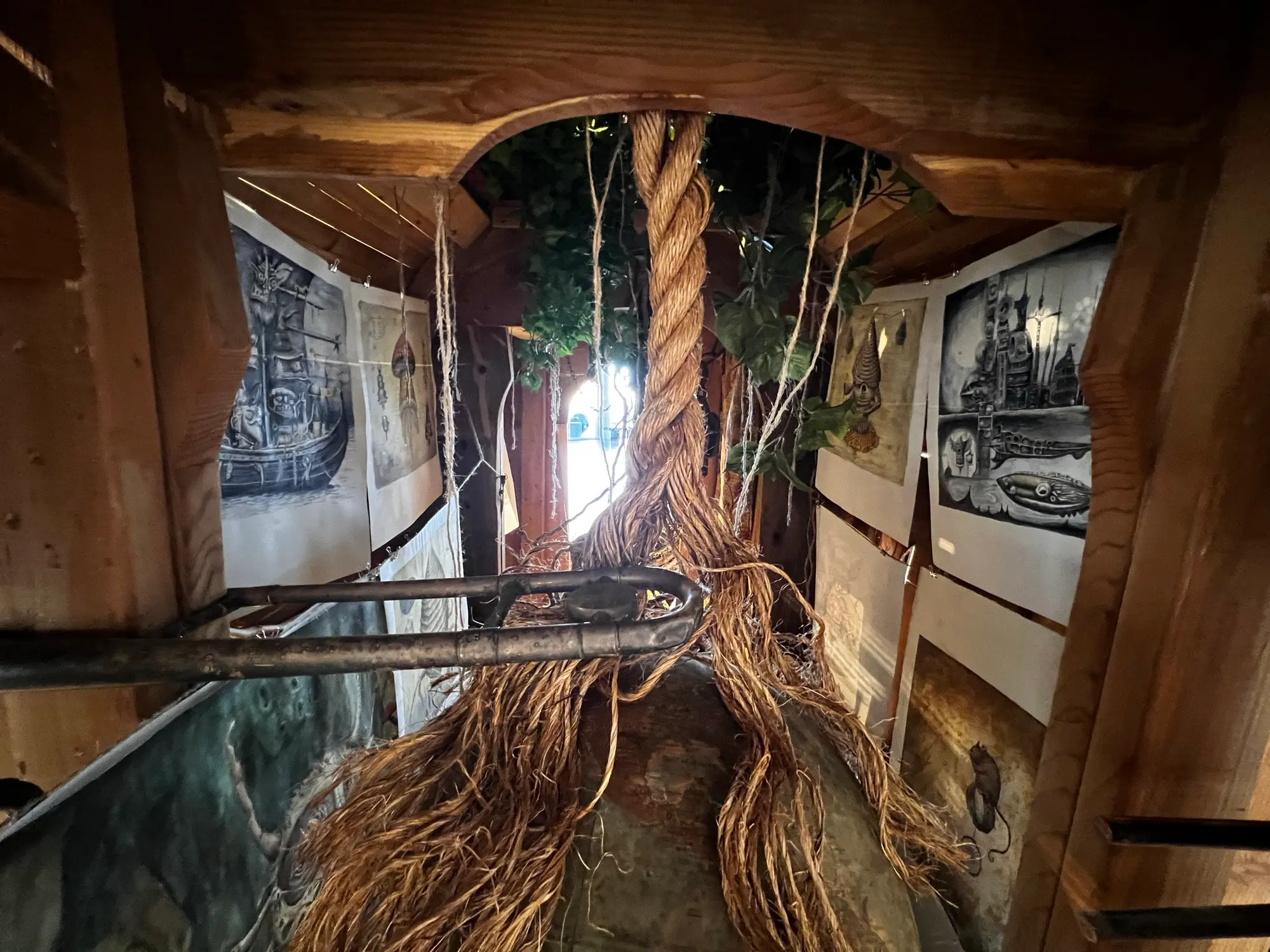 Inside the wagon: a thick rope descends from the ceiling greenery and frays into root strands over a globe, with watercolor studies hung on lines along both walls toward a lit doorway.