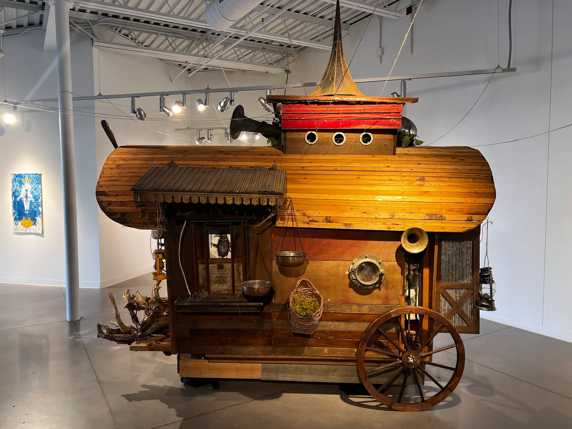 The wagon's far face: a corrugated awning over a shuttered window, hanging scale pans and a moss basket, a bronze porthole, a brass horn, and a large wagon wheel; a blue cyanotype work hangs on the gallery wall beyond.