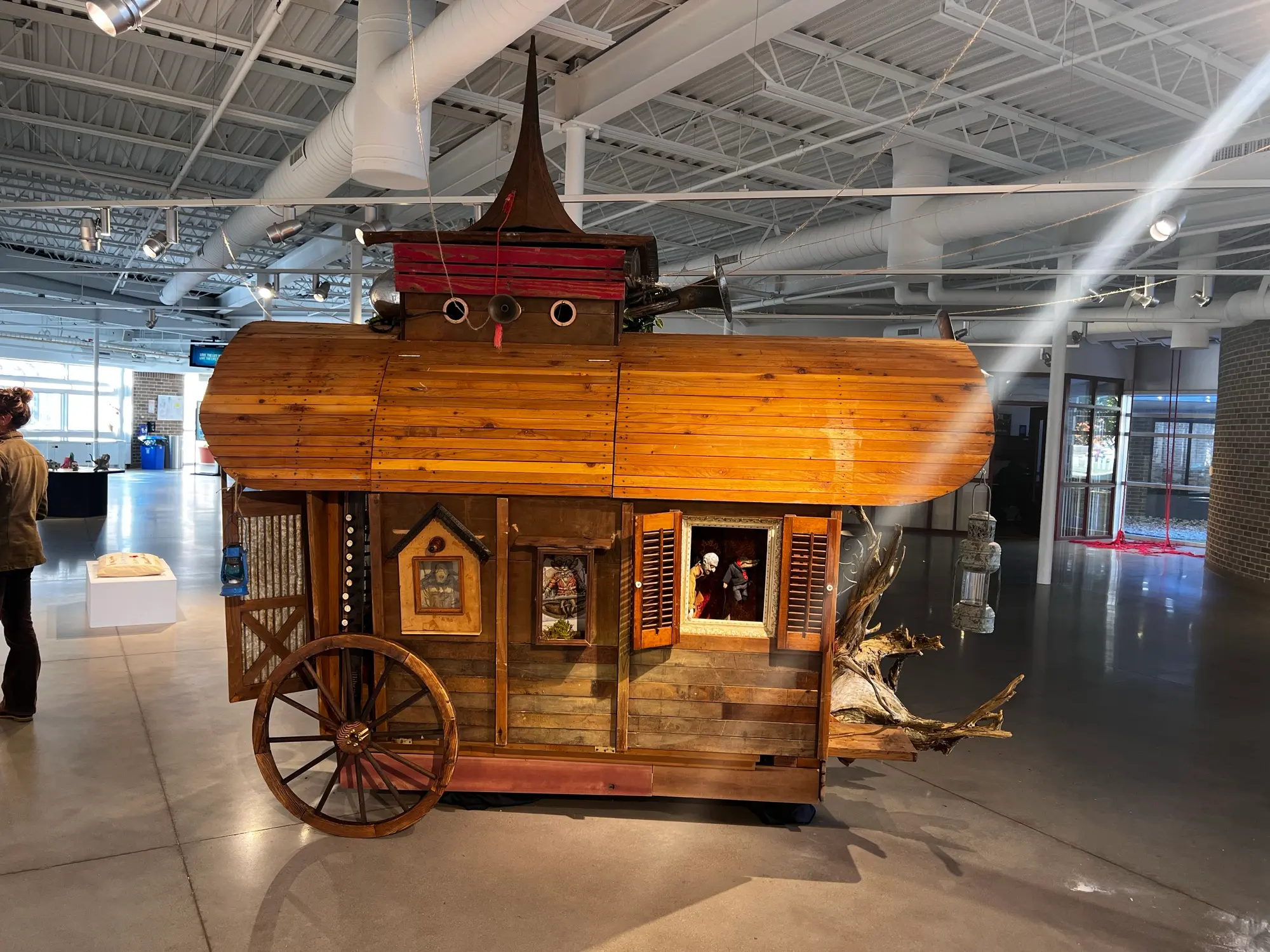 A wooden field-naturalist's wagon on a gallery floor: plank-clad rounded roof, a spired cupola with portholes, shuttered windows with puppets inside, a wagon wheel, lanterns and driftwood roots at one end.